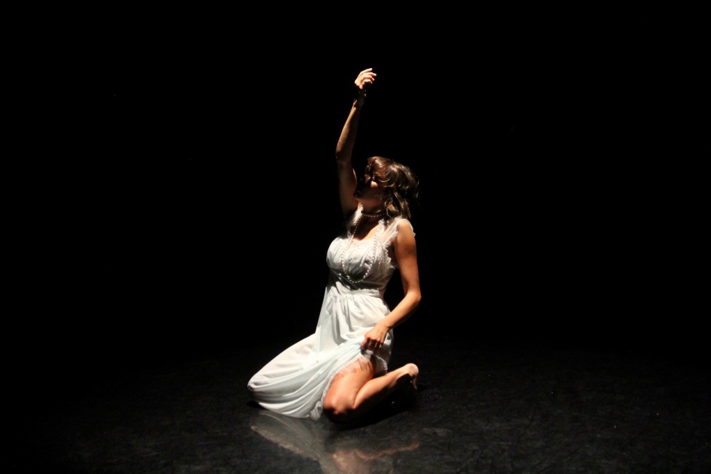 Kneeling on the ground, with left arm raised above head. Dancer wearing blue dress and pearls. 